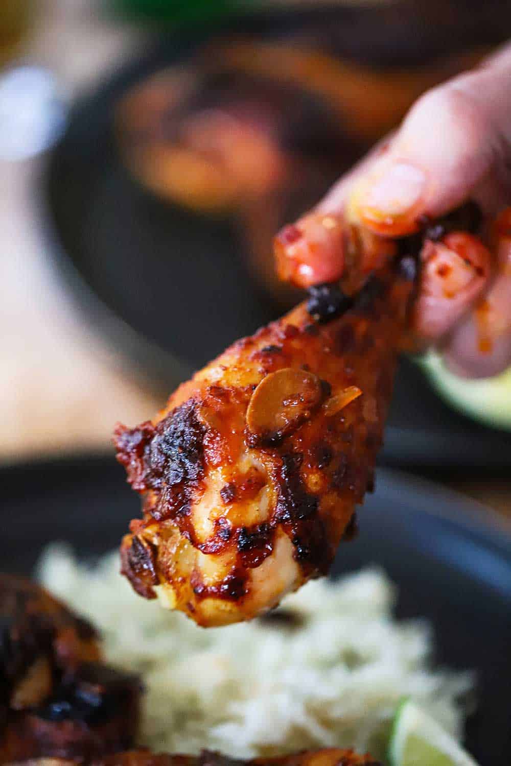 A hand, with a little sauce on the fingers, holding a roasted chipotle chicken leg up over a plate of cilantro-lime rice.