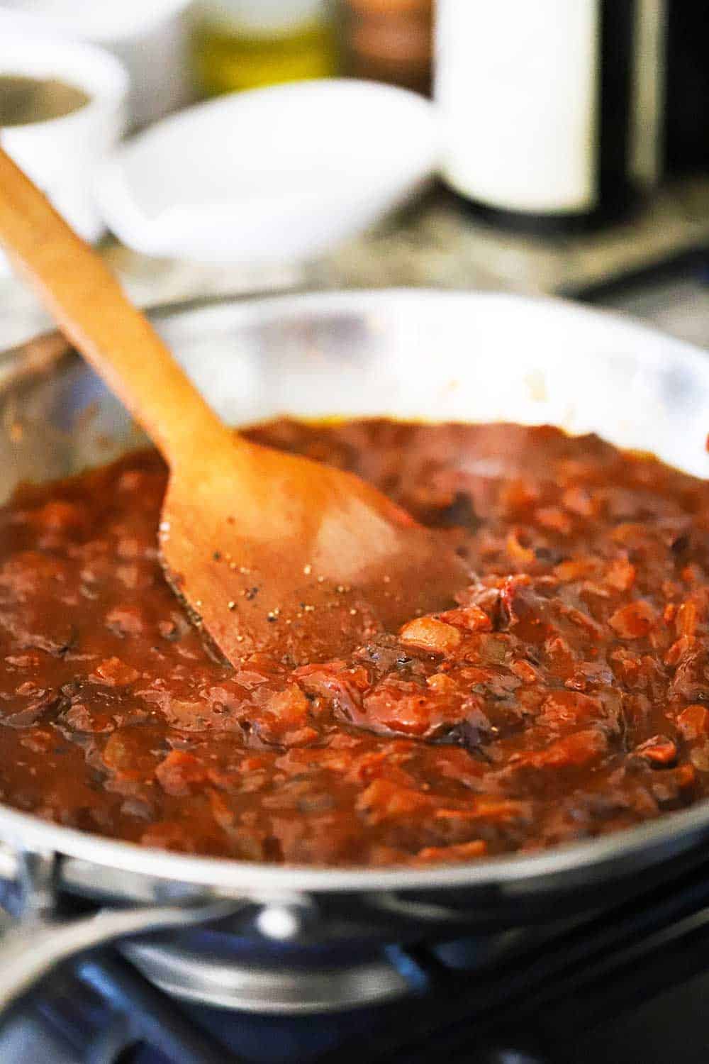 A large stainless steel skillet filled with a chipotle tomato sauce with a wooden spoon in the middle of the simmering sauce.