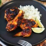 A black dinner plate filled with two pieces of roasted chipotle chicken, cilantro-lime rice, and 2 lime wedges, all sitting next to a glass of Mexican beer.