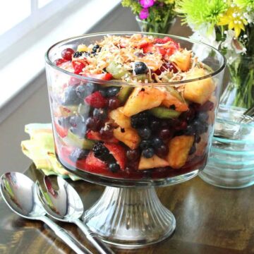 A glass trifle filled with a summer fruit salad topped with toasted coconut sitting on a table near a window.
