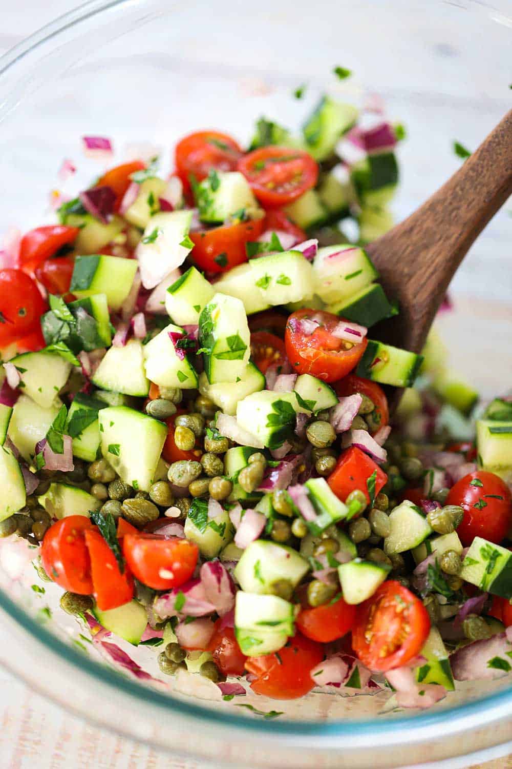 A glass bowl containing a mix of chopped tomatoes, cucumbers, capers, olives, and herbs with a wooden spoon in the middle of it.