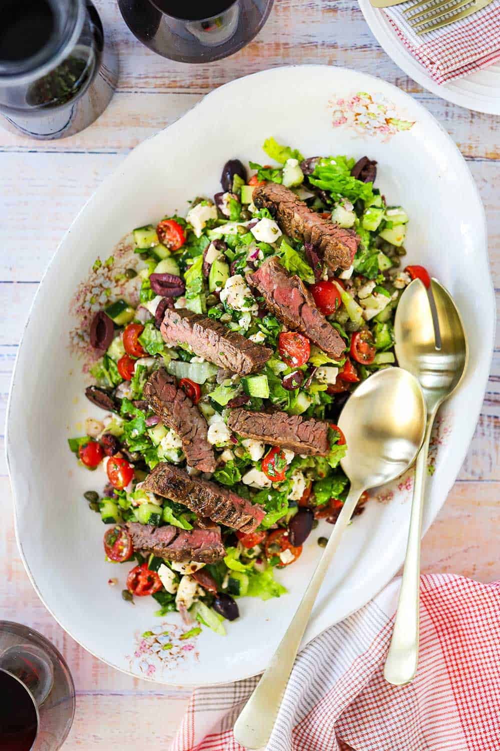 A large oval platter filled with a Greek salad and grilled steak with two large gold spoons on the side.