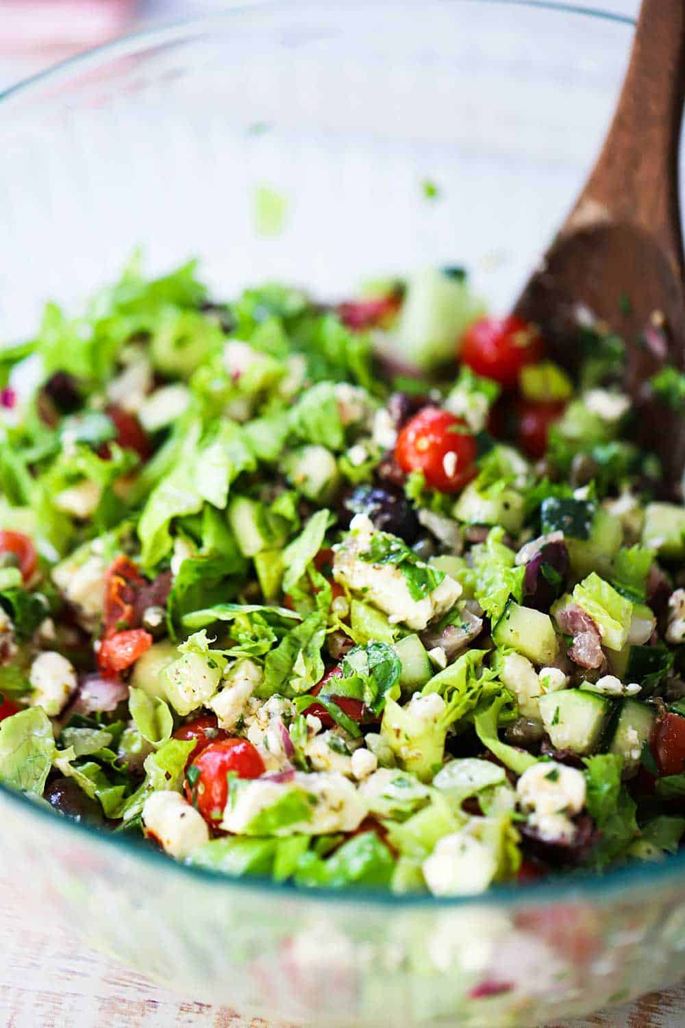 A glass bowl filled with a chopped Greek salad with a wooden spoon stuck in the middle.