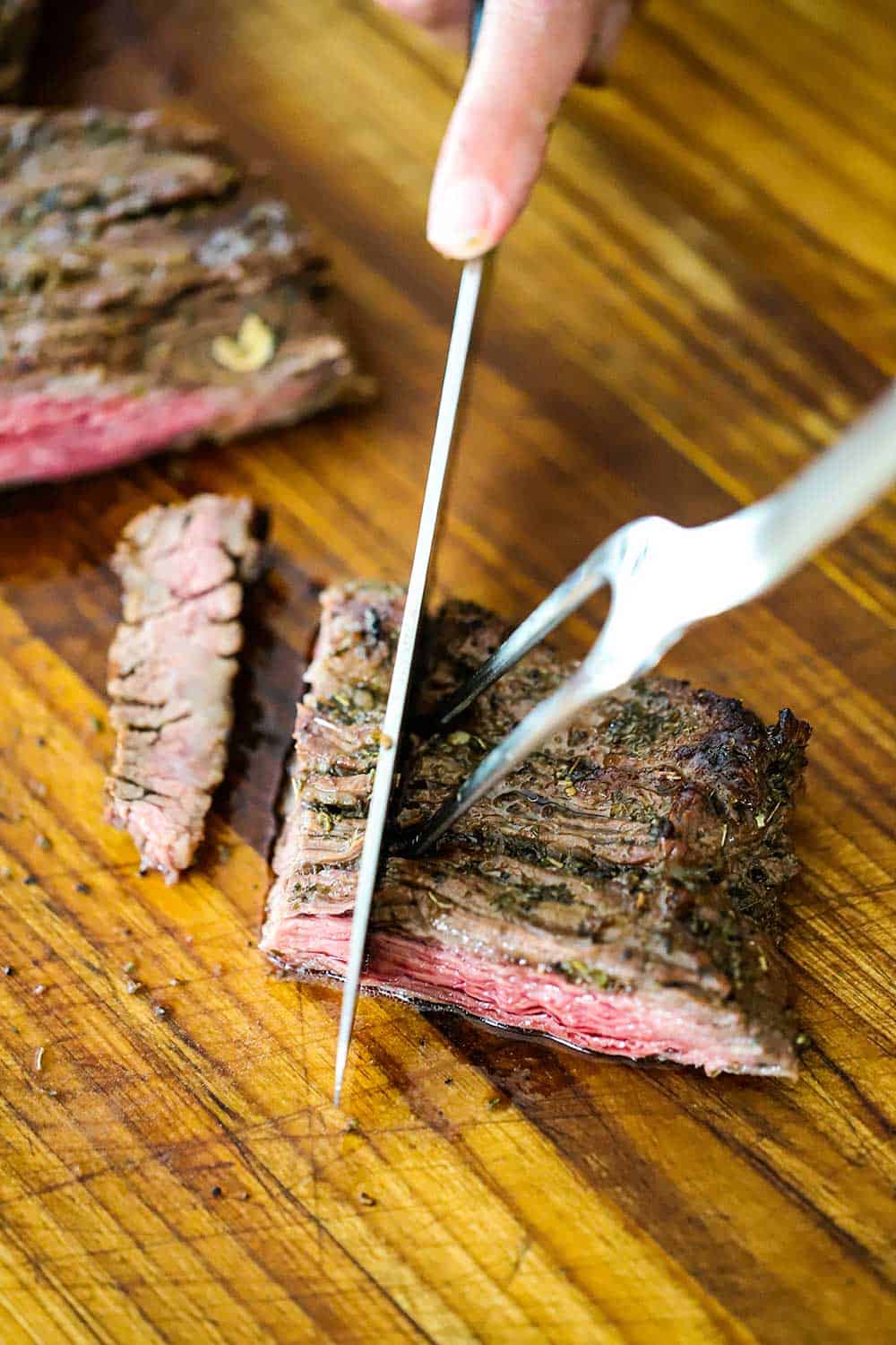 A hand using a carving knife and fork to slice a piece of grilled skirt steak against the grain to form bite-size strips.