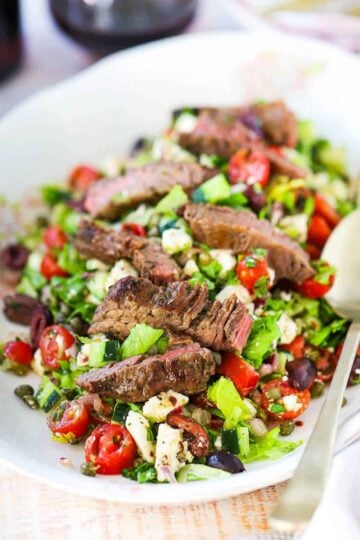 A Greek salad on a large oval platter topped with strips of grilled meat with two gold serving spoons on the side.