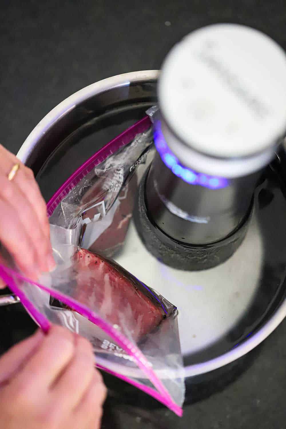 Two hands sealing a baggie filled with a steak that is being submerged into a pot of water with a sous vide device in the water.