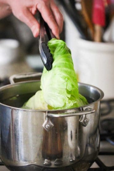A hand using a pair of tongs to life a cabbage leaf out of a pot of boiling water.