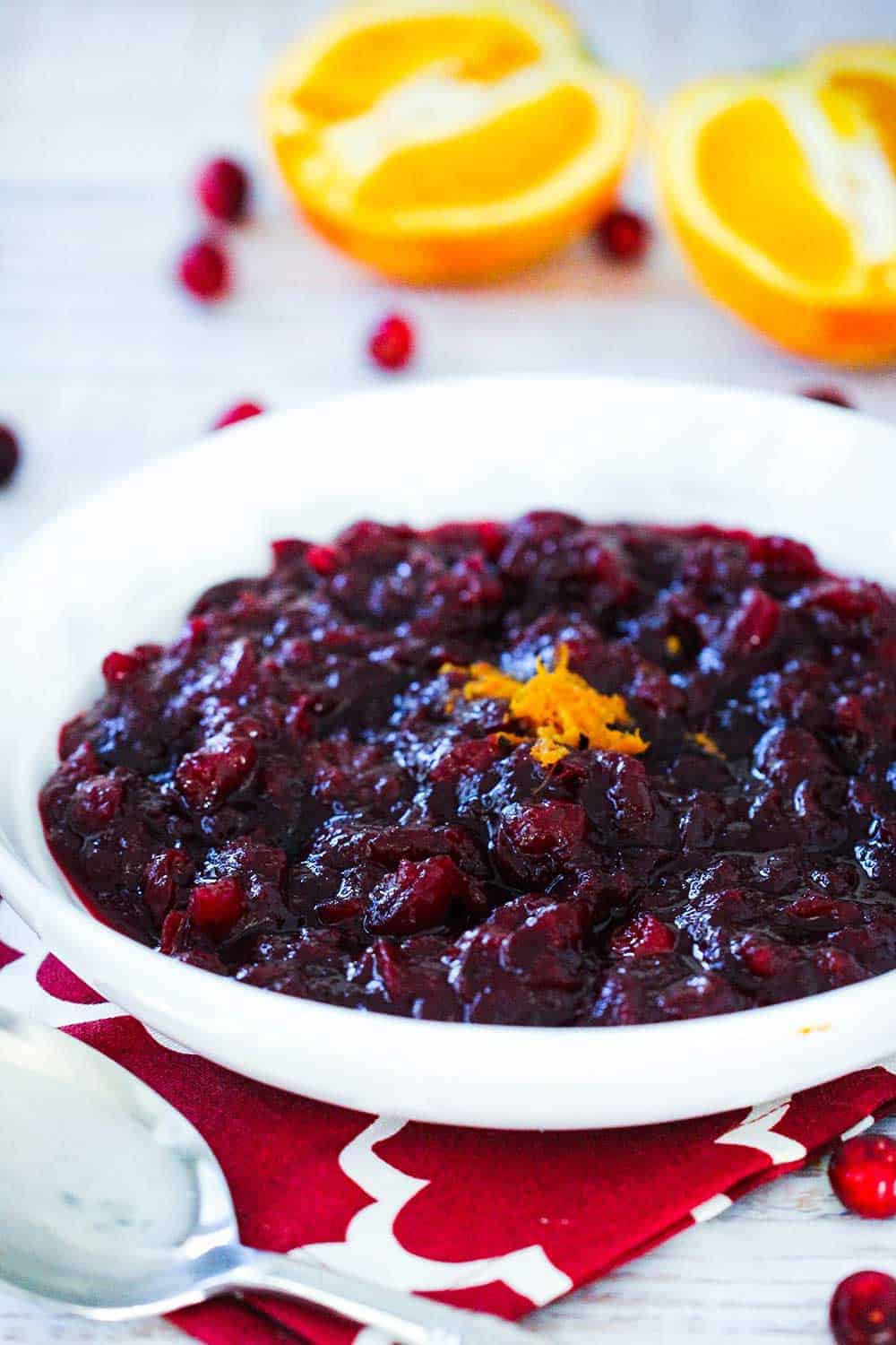 A white bowl containing Maple Orange Cranberry Sauce next to sliced oranges and cranberries.