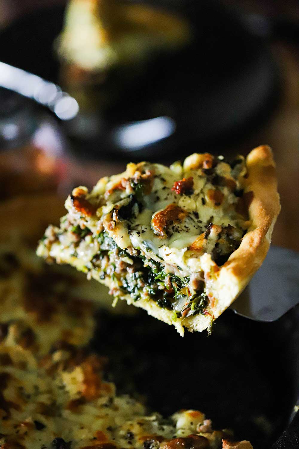 A slice of deep-dish pizza with sausage and spinach being lifted from the pan with a spatula.