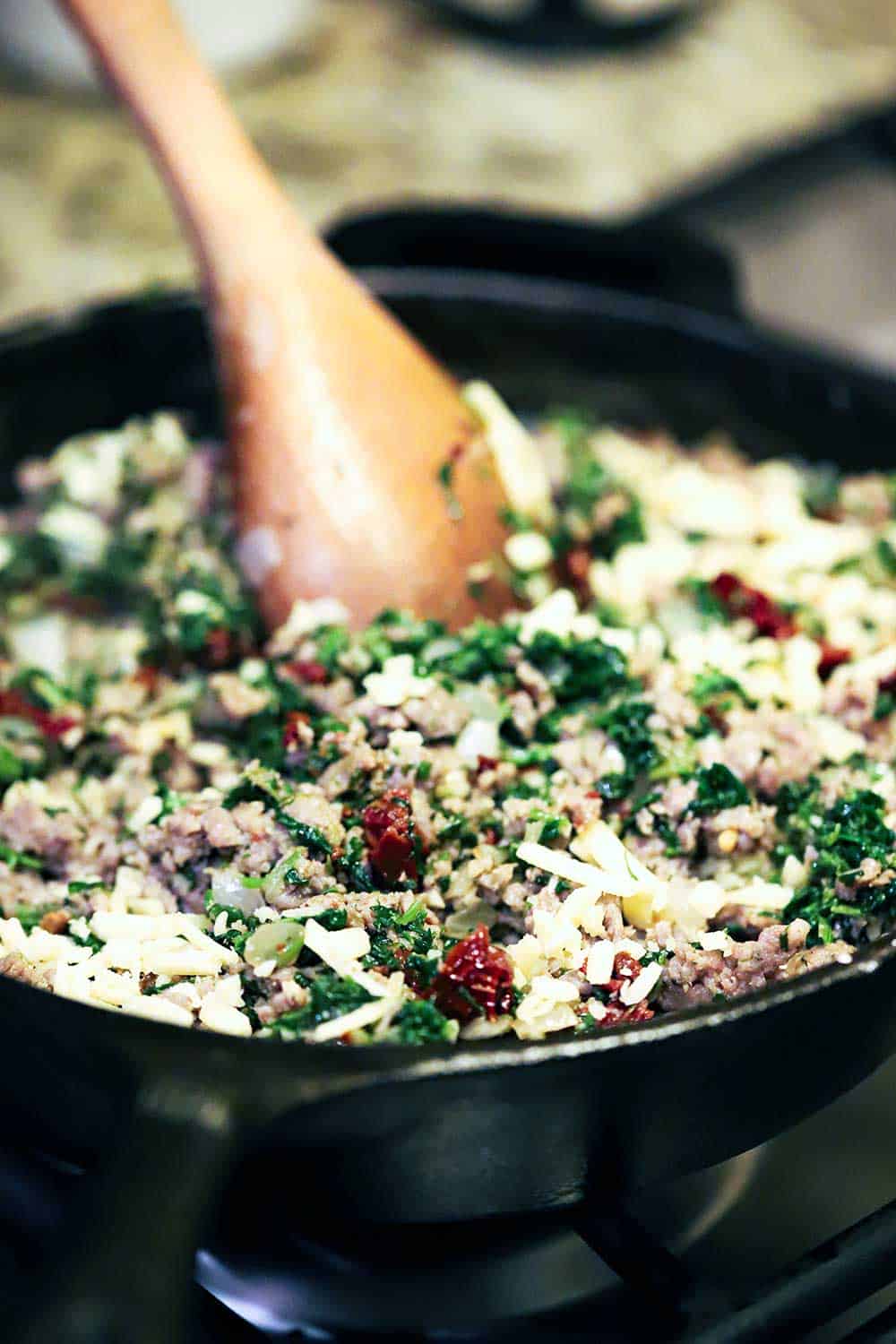A wooden spoon being used to mix cooked sausage, chopped spinach, sun-dried tomatoes, and shredded cheese in a cast-iron skillet.