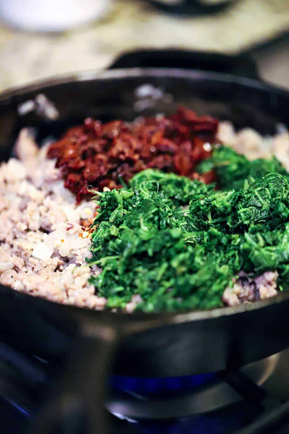A cast-iron skillet filled with cooked Italian sausage with chopped spinach and chopped sun-dried tomatoes sitting on top of the meat.