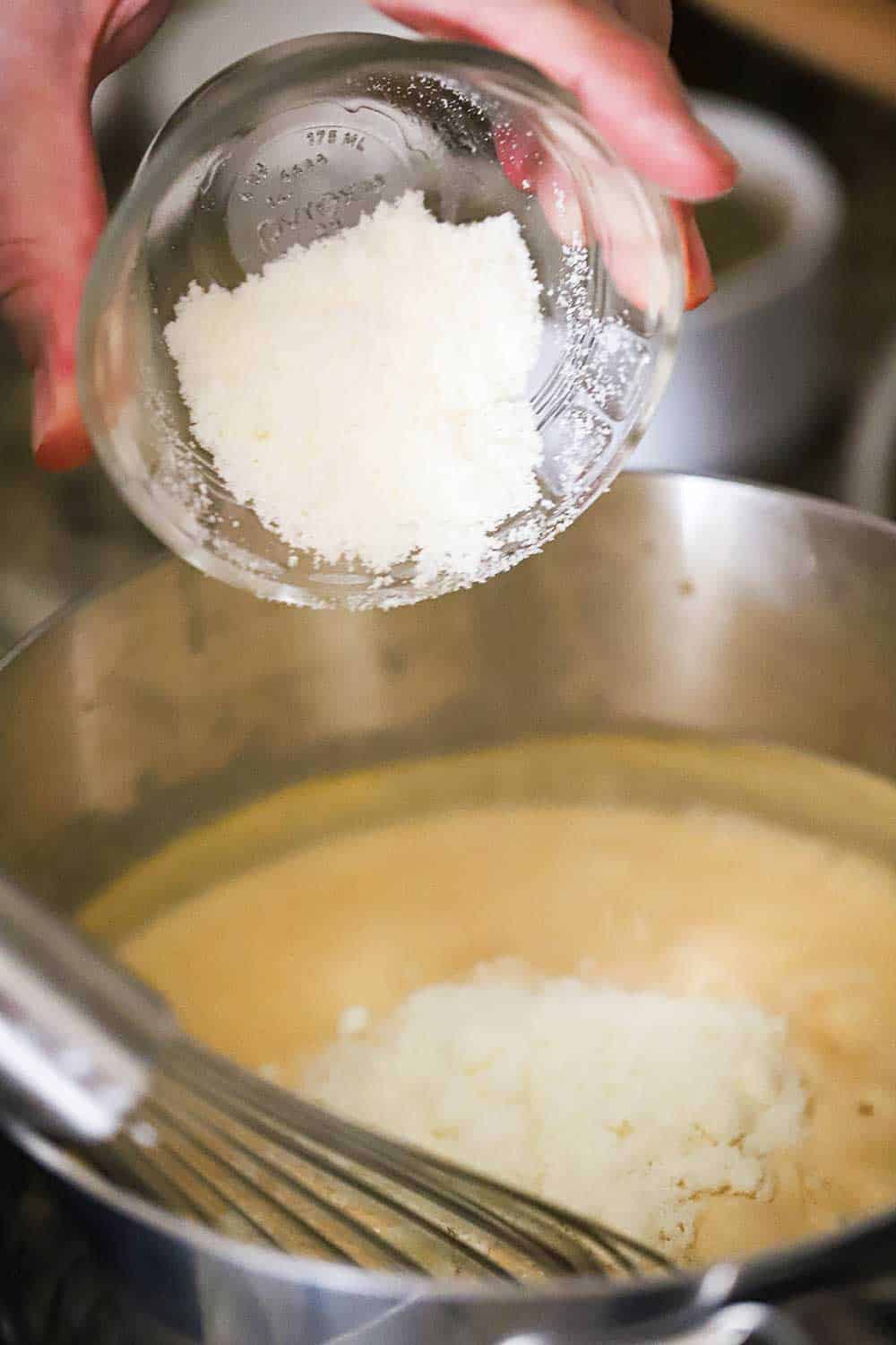 A hand dumping ¼ cup of grated Parmesan cheese into a saucepan filled with a bechamel sauce.