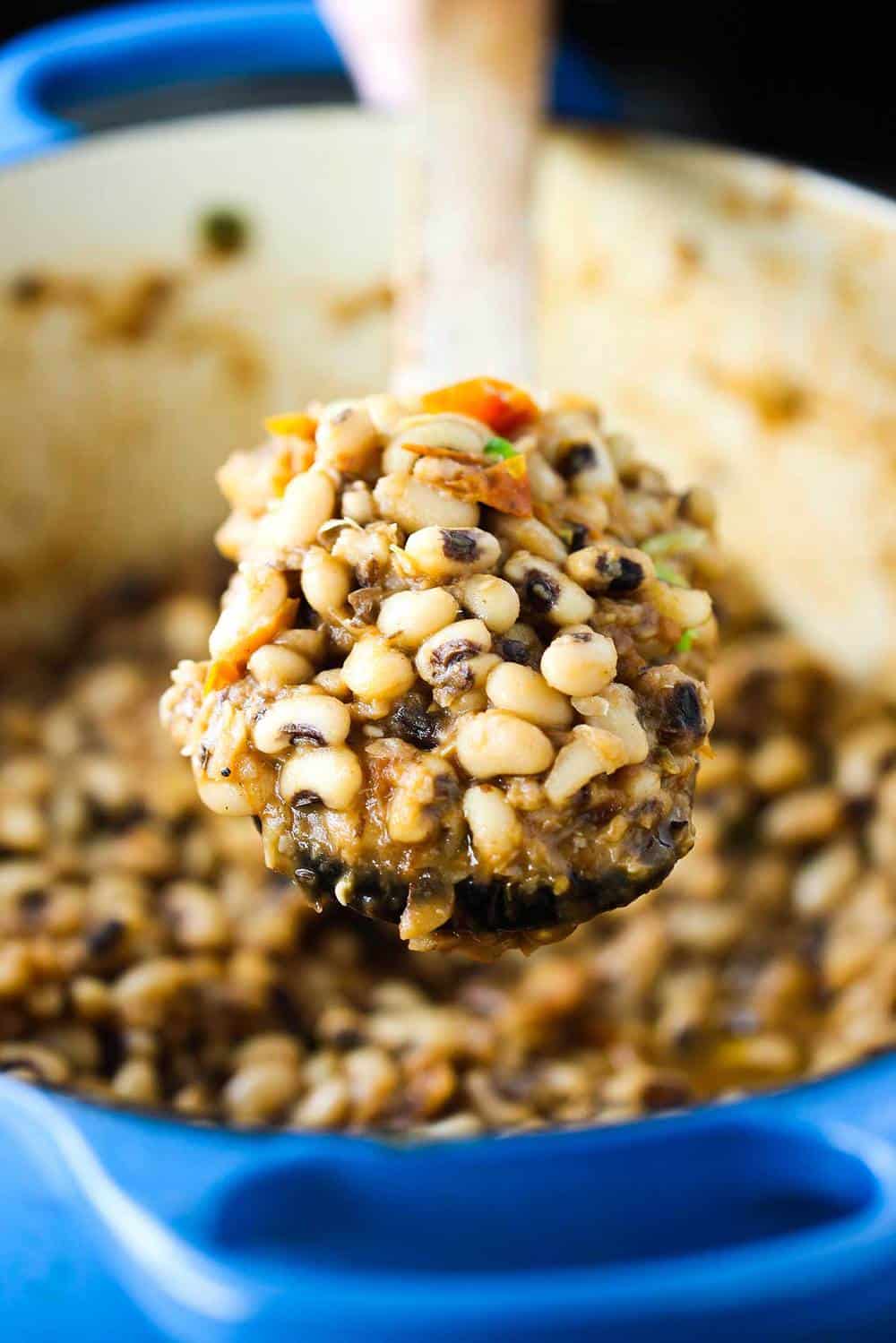 A wooden spoon full of black-eyed peas with tomatoes in a large blue pot.