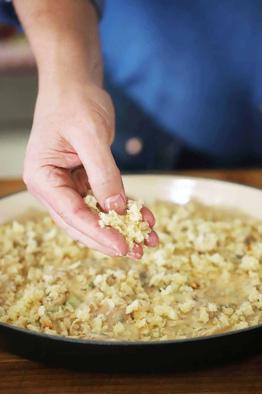 A person sprinkling fresh bread crumbs over the top of an uncooked oyster casserole in a black oval baking dish.