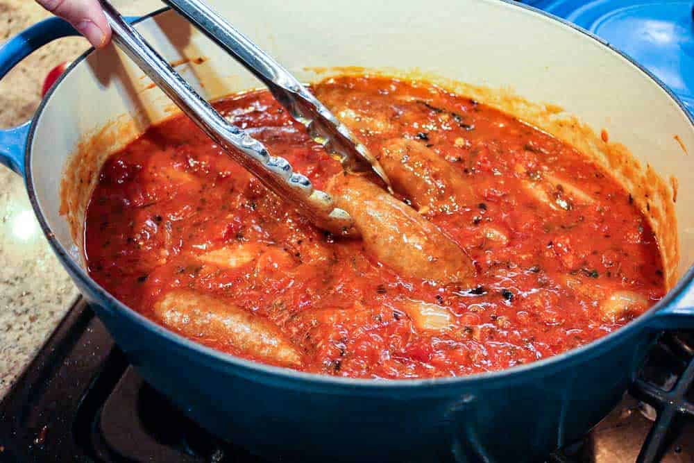 A pair of tongs lowering an Italian sausage into simmering marinara sauce