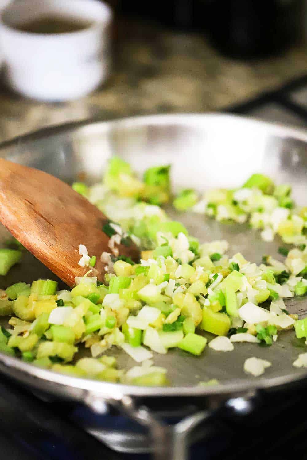 Onions, celery, and green onions being sautéed in a large silver skillet with a wooden spoon in the middle of it.