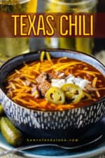 A large blue bowl filled with Texas chili topped with cheese, onions, and jalapenos next to a glass of beer and a jar of pickles.