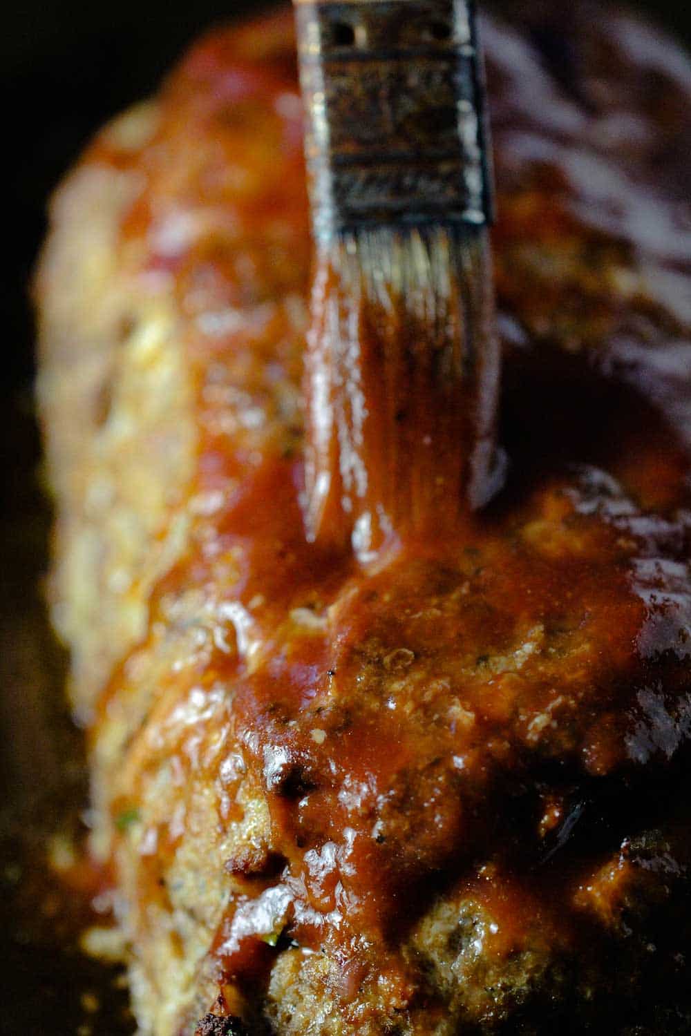 A hand using a brush to apply a tomato sauce to the top of the best-ever meatloaf, partially cooked.