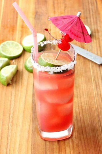 A tall glass holding a Mexican Sea Breeze Cocktail with a pink straw on a wood cutting board with lime slices