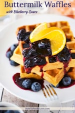 A stack of buttermilk waffles topped with a blueberry sauce and a slice or orange all on a white plate with a fork next to it.