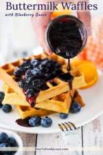 A hand pouring a blueberry sauce over a stack of buttermilk waffles on a white plate next to a fork.