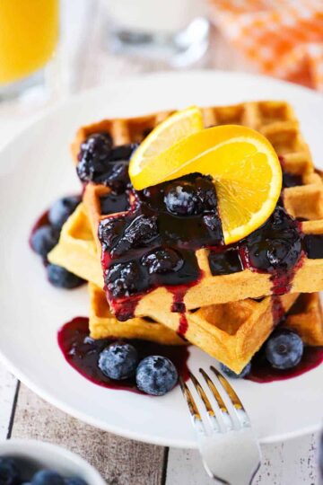 A stack of buttermilk waffles topped with a blueberry sauce and a slice or orange all on a white plate with a fork next to it.