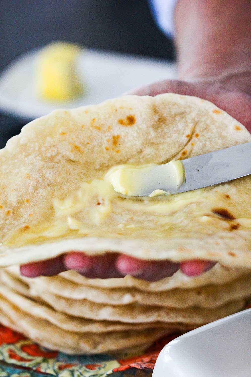 A knife spreading butter onto a warm homemade flour tortilla.