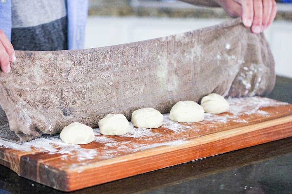 A damp kitchen towel being lowered over flour dough balls on a cutting board.