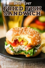 A straight-on view of a fried fish sandwich with a bite taken out of it sitting on a black dinner plate and a glass of beer in the background.