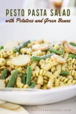 A close up view of a large circular white serving bowl filled with pesto pasta salad with sliced potatoes and green beans.