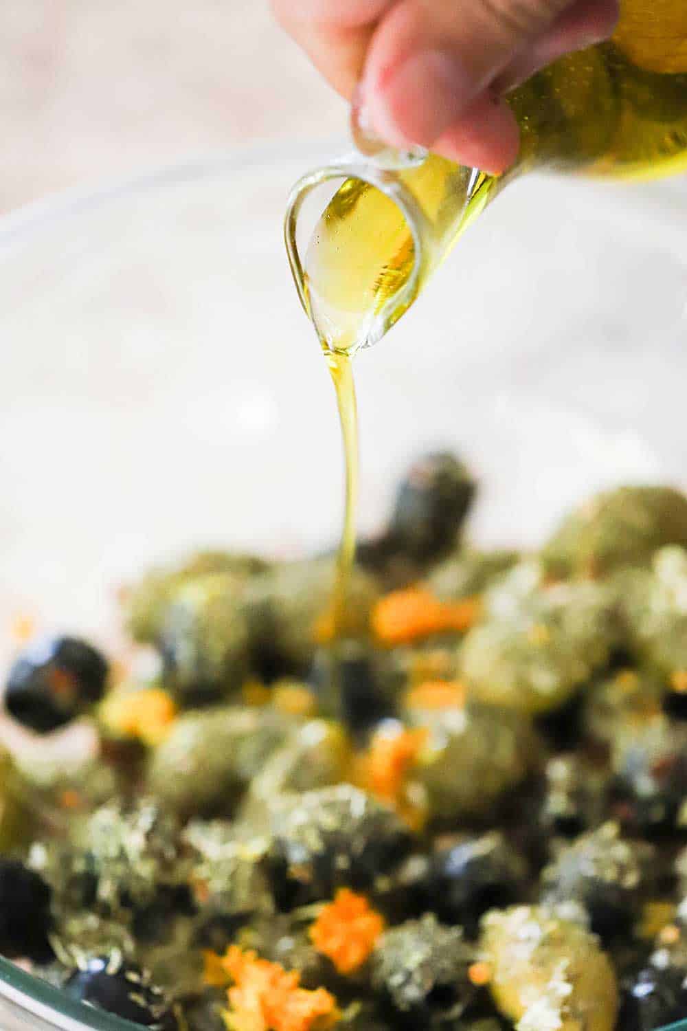 A hand pouring olive oil from a glass oil holder into a glass bowl filled with marinated olives.