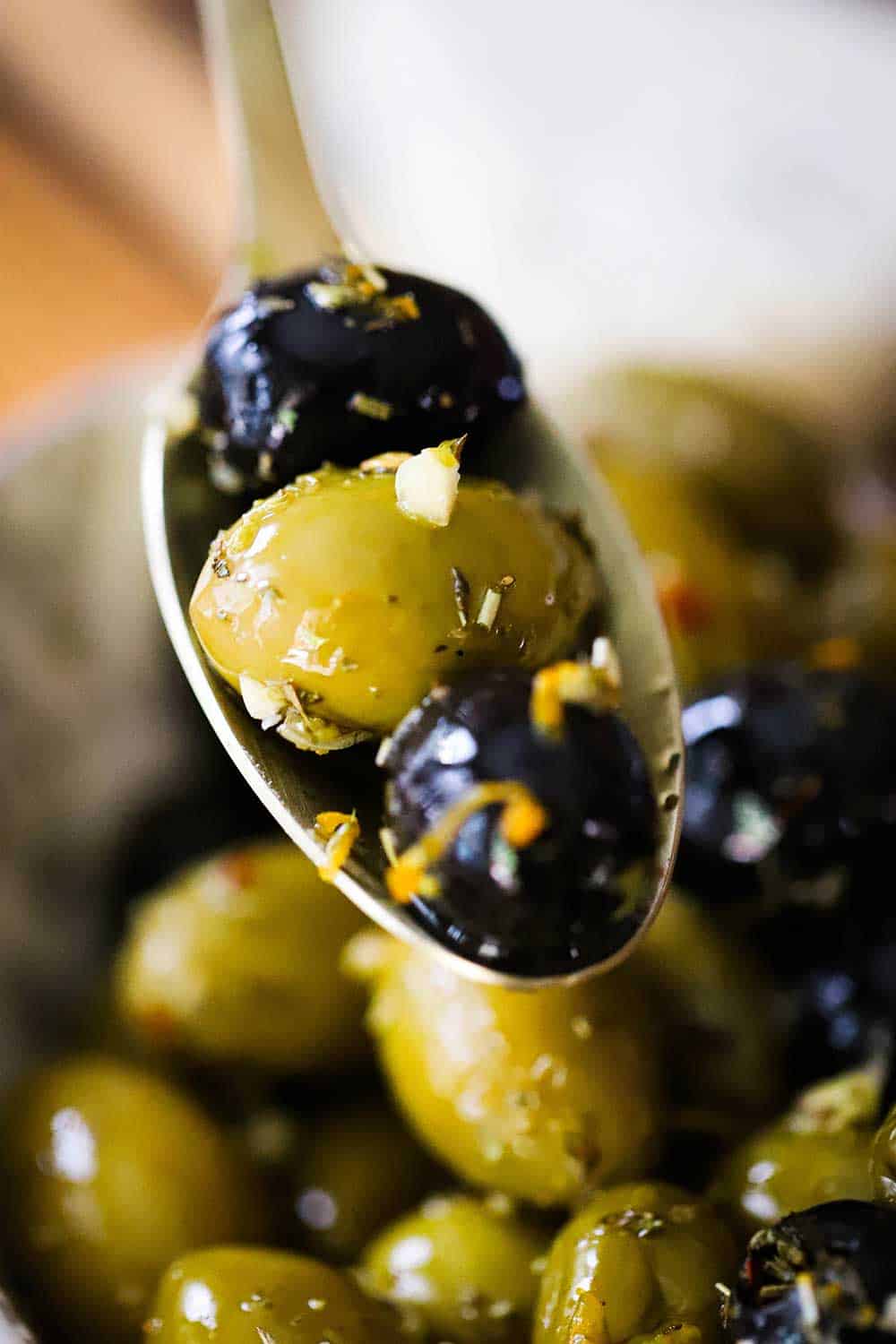 A spoon holding up three marinated olives over a bowl of the same.