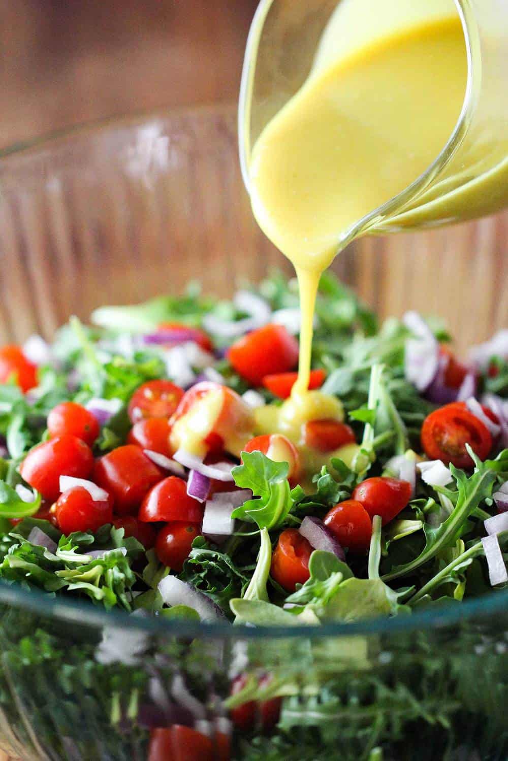 Arugula salad with tomatoes in a glass bowl with vinaigrette being poured over it.
