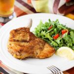 Pork Milanese on a white plate next to an arugula salad and lemon wedge.