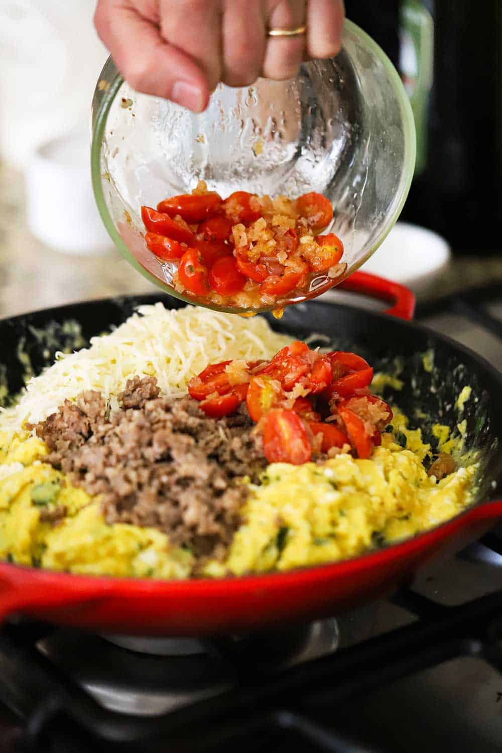 A hand transferring sautéd tomatoes from a small glass bowl into a large skillet filled with scrambled eggs, cooked sausage, and shredded mozzarella cheese.