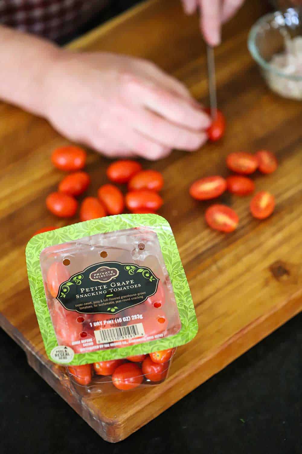 A person using a small knife to slice grape tomatoes in half on a cutting board in front of a package of the tomatoes.