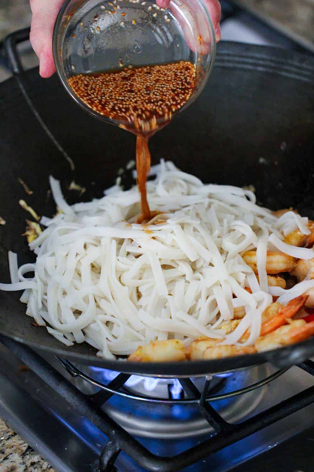 A hand pouring pad Thai sauce into a wok filled with rice noodles.