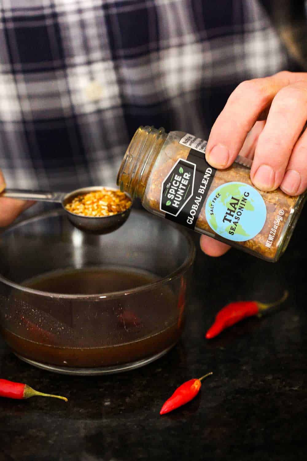 A hand pouring Thai seasoning into a tablespoon over a glass bowl of pad Thai sauce.