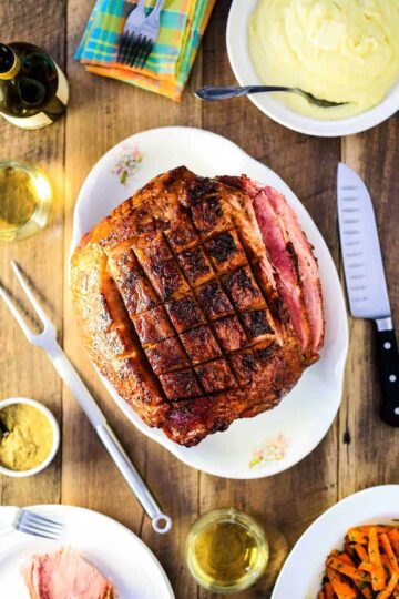 An overview shot of a honey bourbon glazed ham next to a bowl of mashed potatoes, glazed carrots, and a glass of white wine.