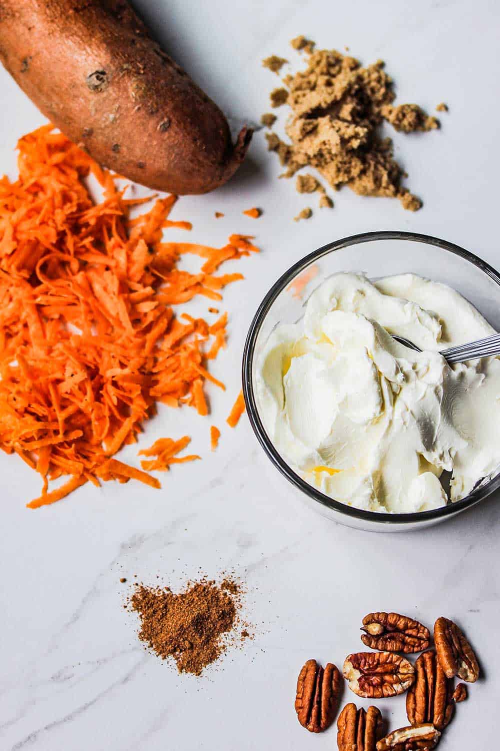 Shredded sweet potatoes next to a whole sweet potato and bowl of cream cheese and pecans.