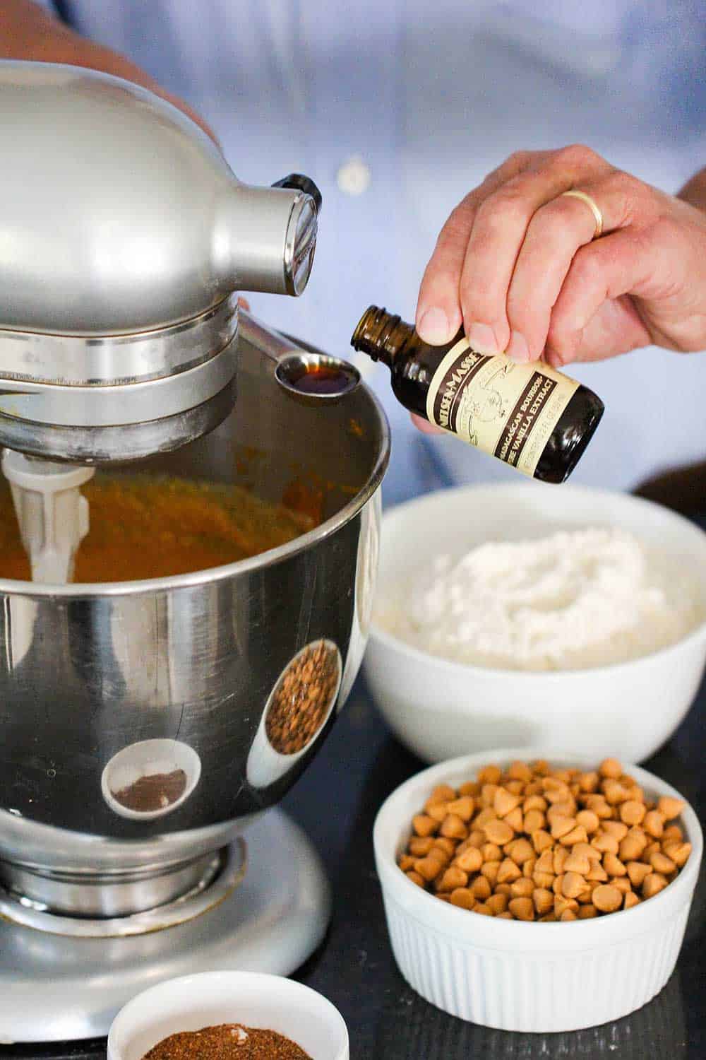A hand adding vanilla extract into a stand mixer with pumpkin bread batter ingredients next to it.