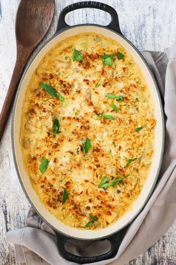 A baked oyster casserole in an oval black baking dish next to a grey napkin and a wooden spoon.