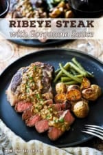 A straight-on view of a ribeye steak that has about half of it cut into sliced and is topped with a creamy gorgonzola sauce and served with roasted potatoes and green beans.