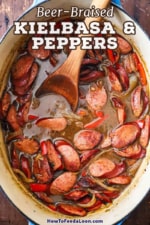 An overhead view of an oval Dutch oven filled with beer-braised kielbasa and onions with a large wooden spoon inserted into it.