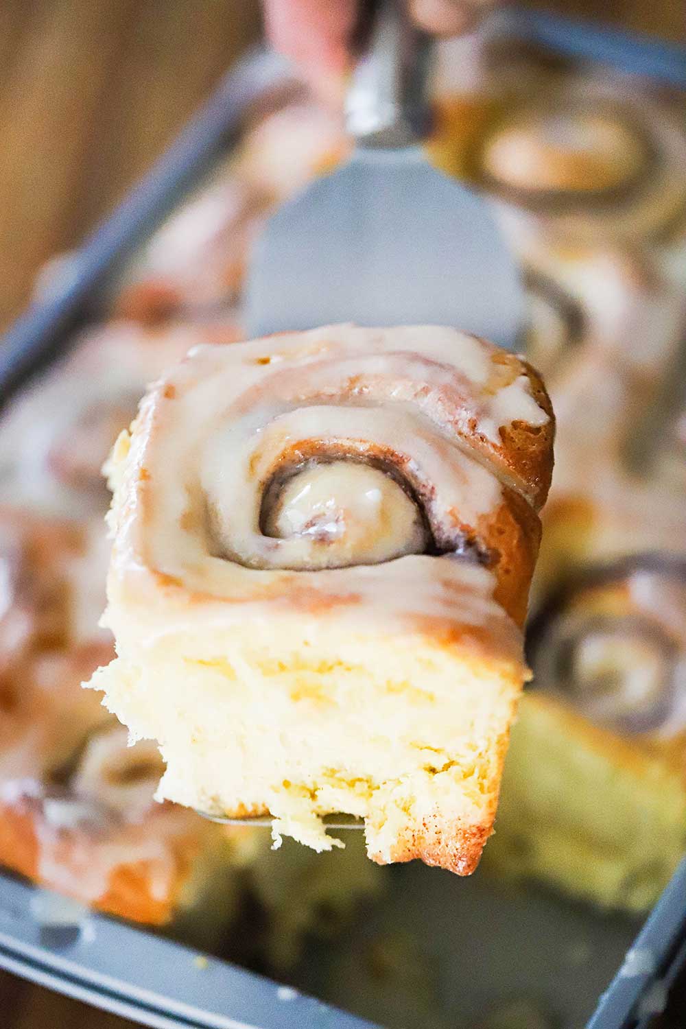 A hand holding a spatula with a homemade cinnamon roll on it over a pan of the rolls.