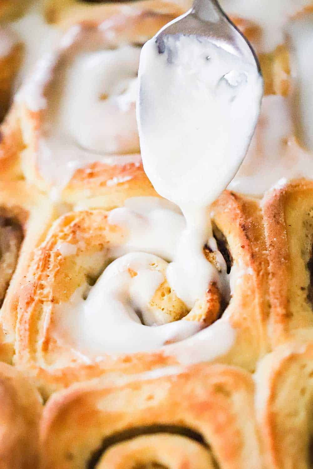 A spoon drizzling icing over the tops of freshly baked cinnamon rolls in a baking pan.