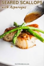 A spoon pouring a brown sauce onto several seared scallops topped with sautéed asparagus.