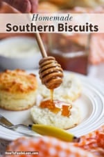 A person using a honey stick to drizzle golden honey over a Southern biscuit that has been split open on a plate.