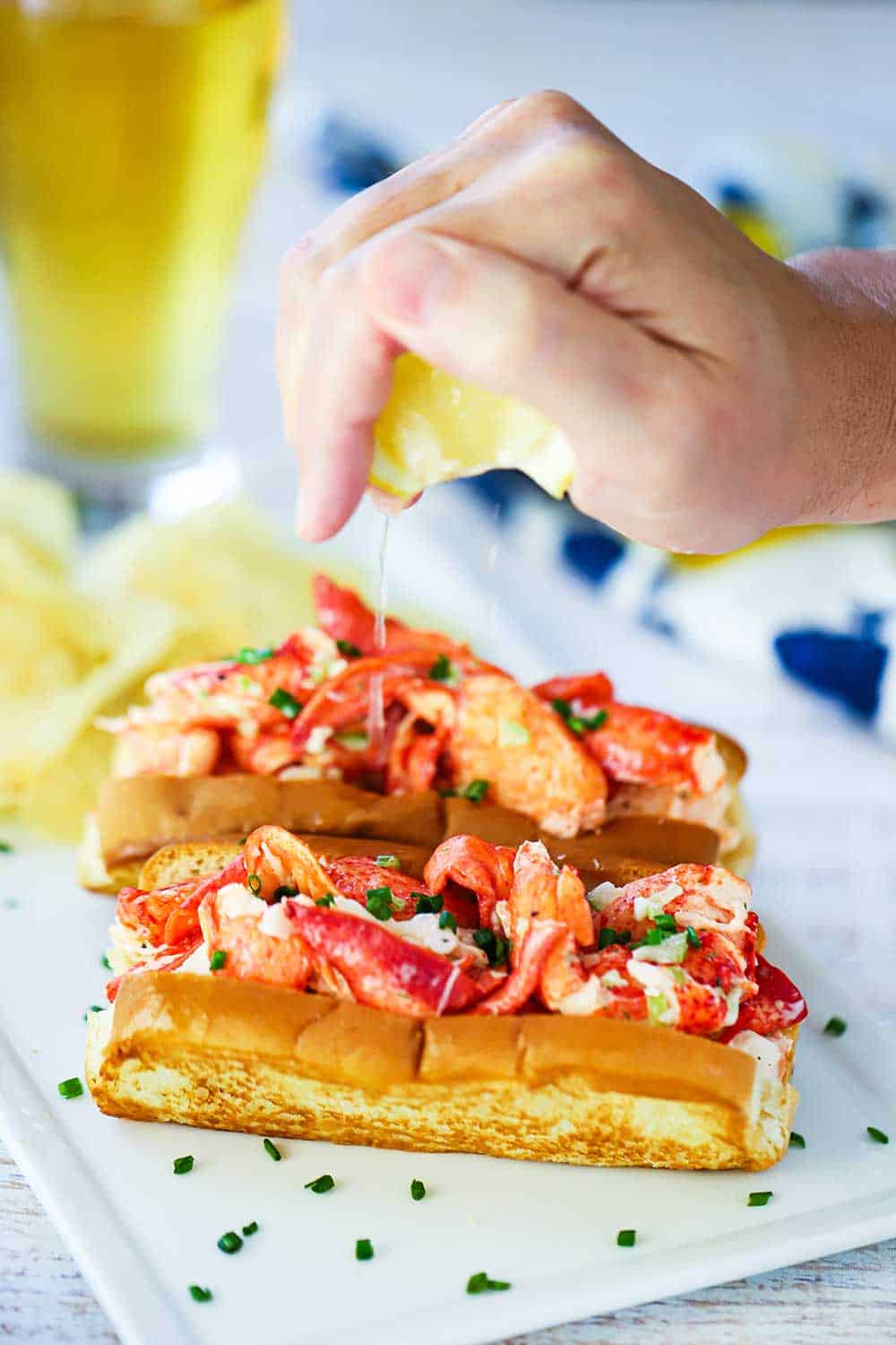 A hand squeezing a lemon wedge over the tops of two lobster rolls on a white plate.