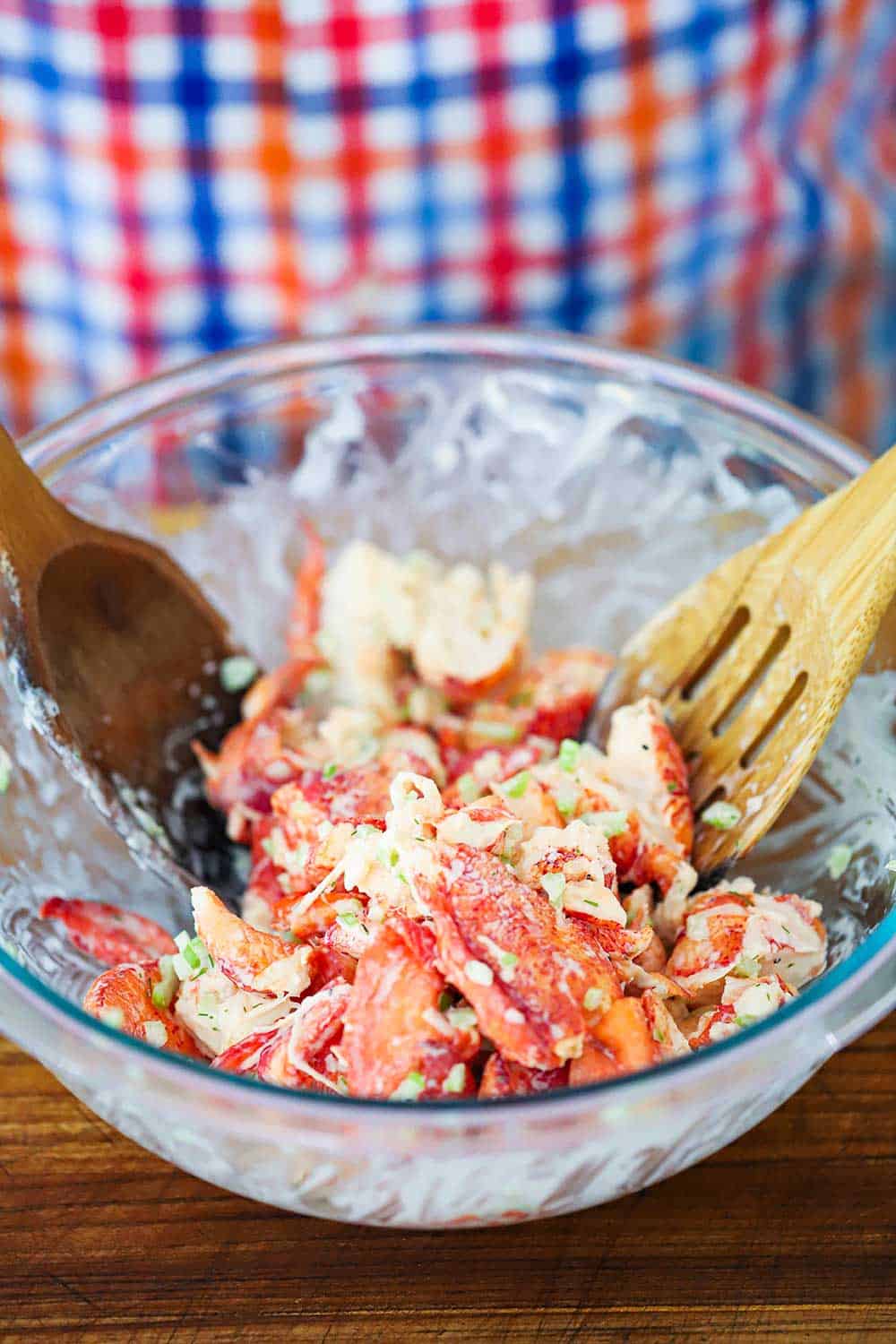 A glass bowl filled with lobster salad that is being tossed by a couple of wooden spoons.